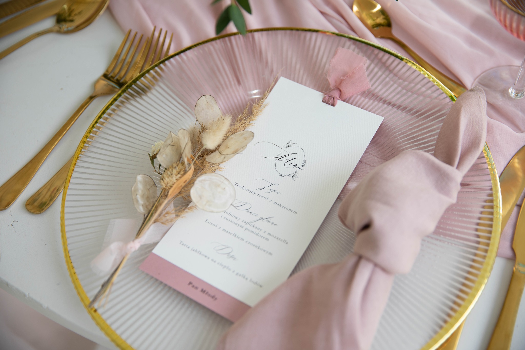 Elegancko nakryty stół z menu weselnym, szklany talerz ze złotym brzegiem, lniana serwetka w pudrowym różu, złote sztućce, suszone kwiaty jako dekoracja.