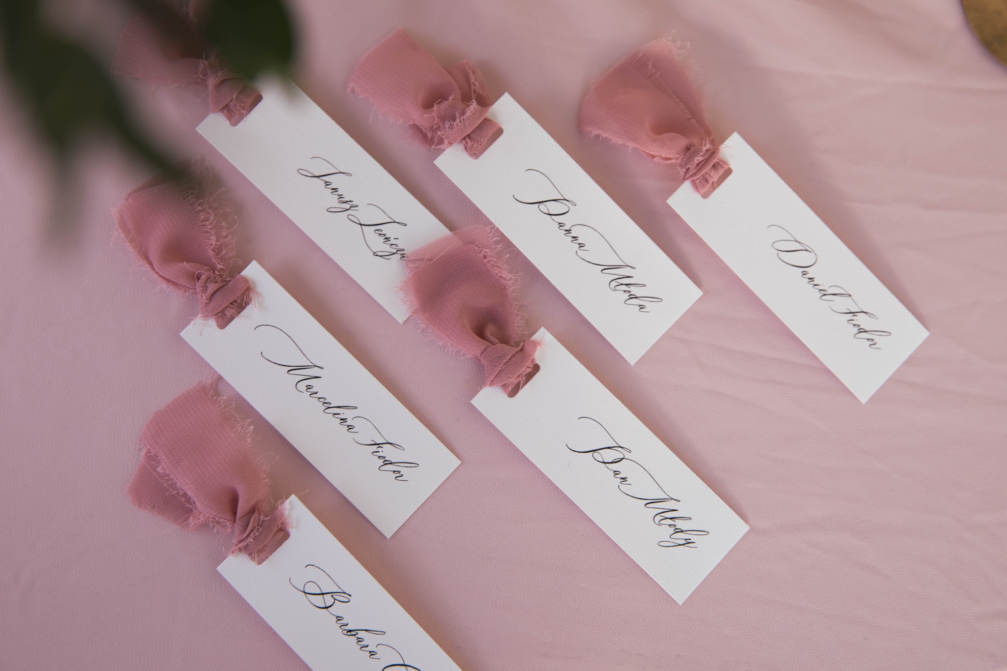 Eleganckie winietki z imionami gości, ozdobione pudrowo-różowymi wstążkami, leżące na delikatnym, różowym tle.