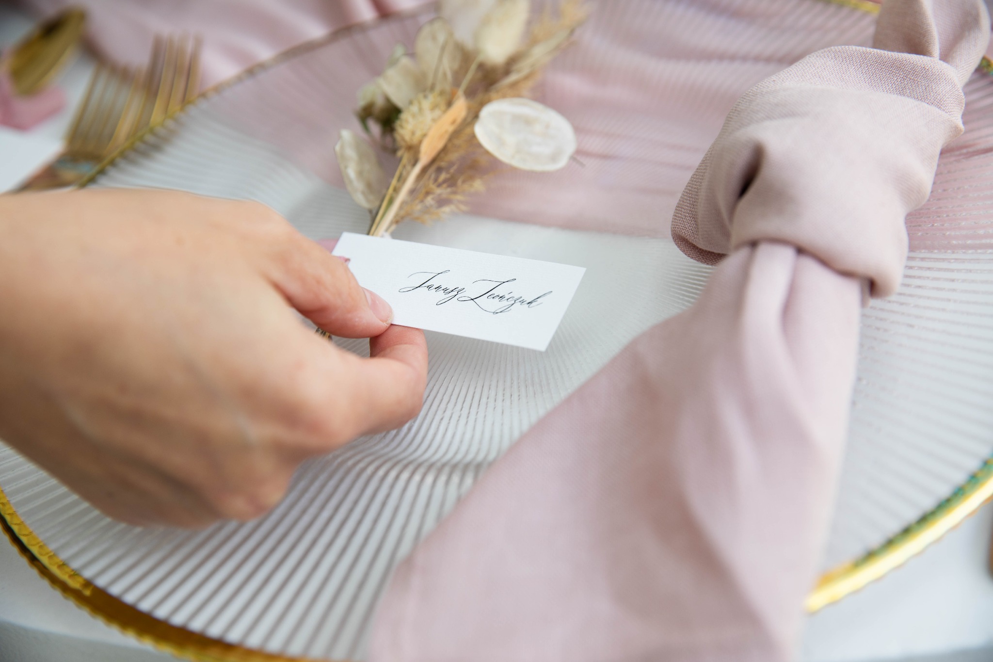 Dekoracja stołu z ręcznie pisaną winietką z imieniem 'Jarosz Leonczuk', suszonymi kwiatami i różową serwetką na szklanym talerzu.
