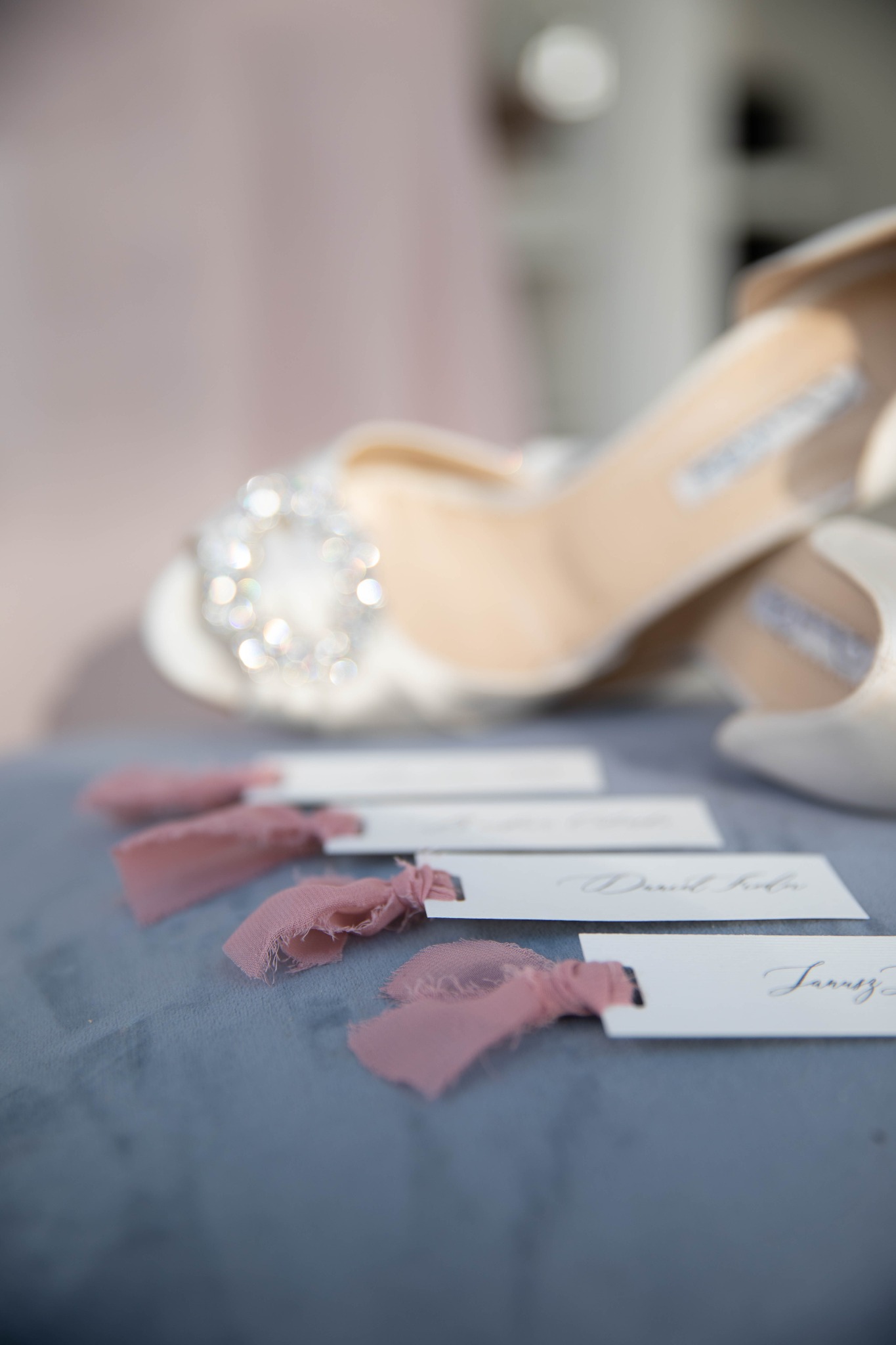 Eleganckie buty ślubne z kryształową ozdobą obok wizytówek z imionami gości, przewiązanych różowymi wstążkami na szarym tle.