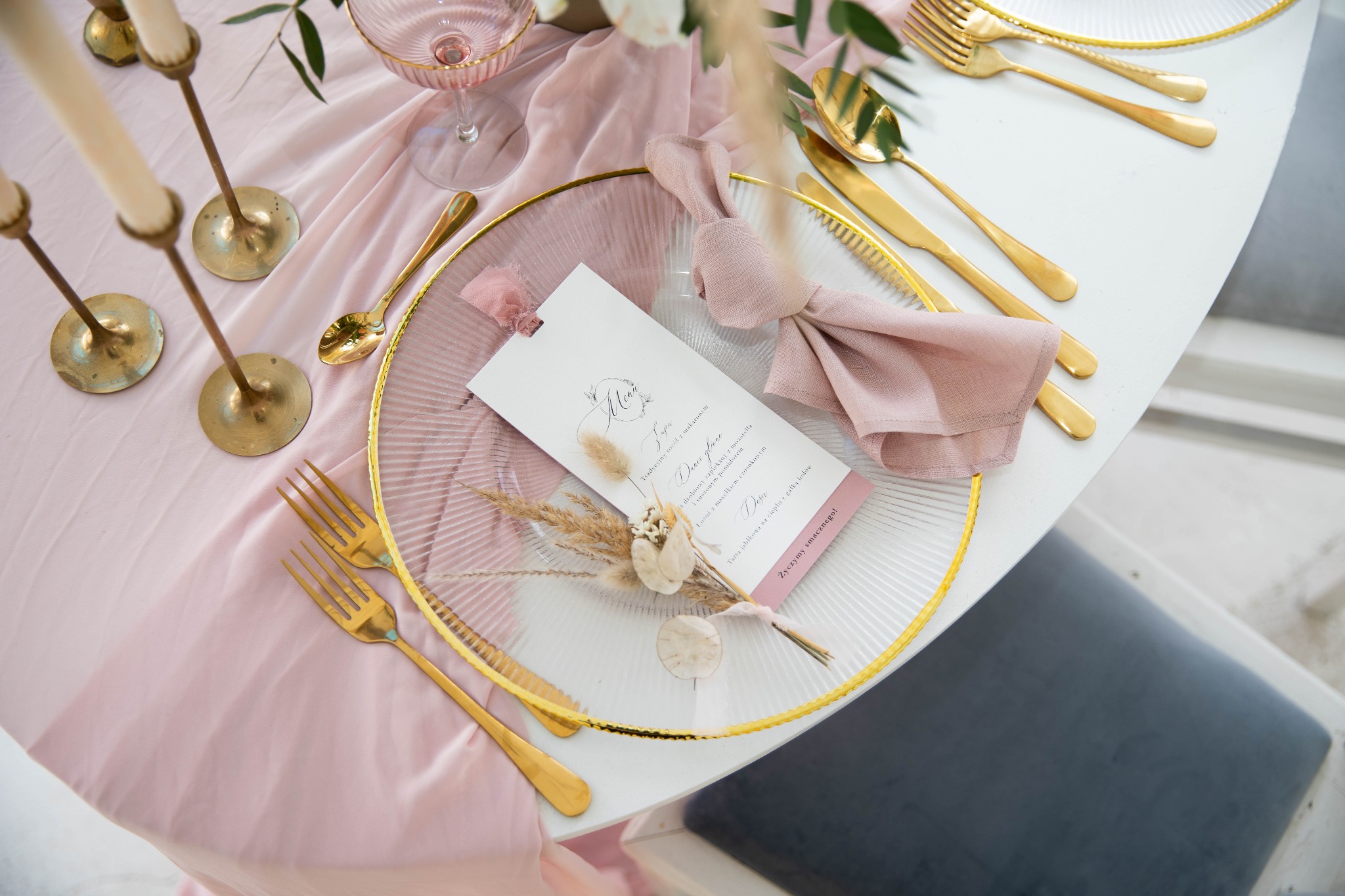 Elegancko nakryty stół z różową zastawą, złotymi sztućcami, szklanym talerzem z menu i dekoracją z suszonych kwiatów, świeczniki w tle.