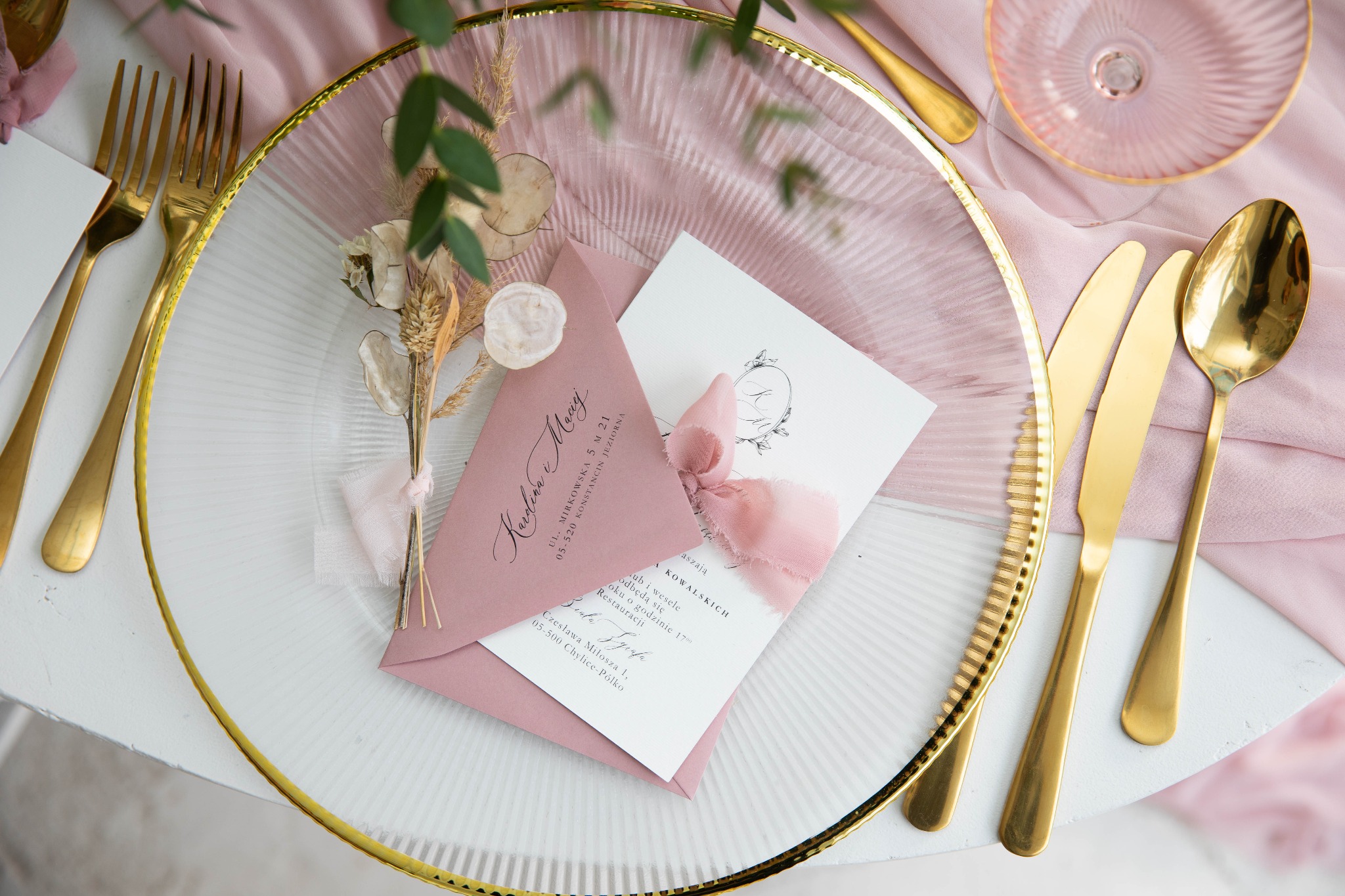 Elegancki stół z przezroczystym talerzem ze złotym brzegiem, złotymi sztućcami, różowym obrusem i zaproszeniem ślubnym w różowej kopercie, przewiązanym różową wstążką.