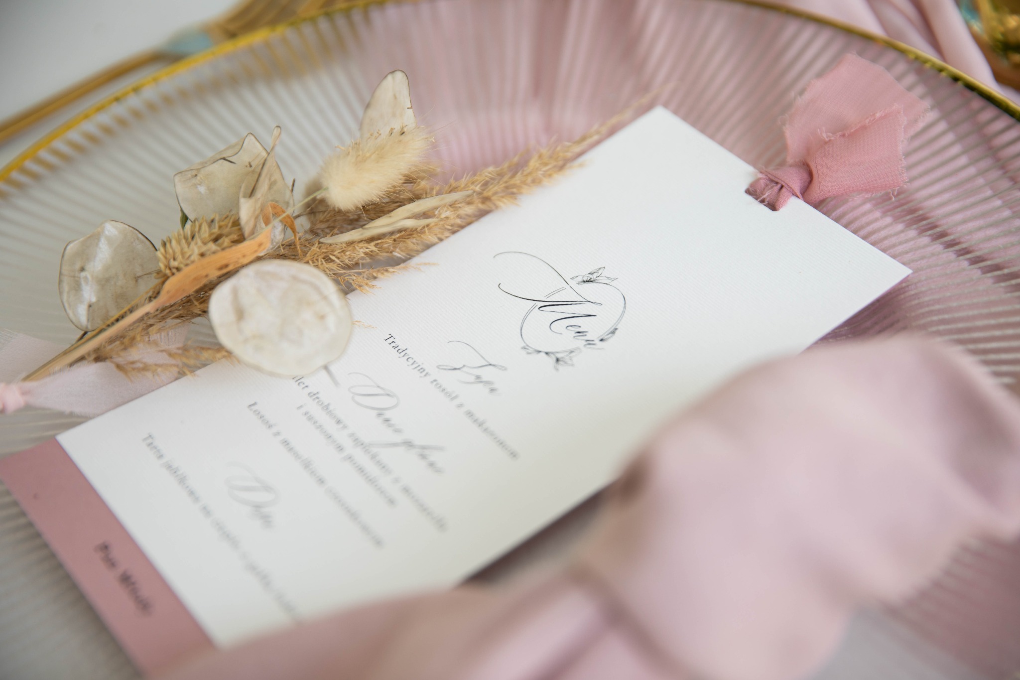 Eleganckie menu weselne w odcieniach bieli i pudrowego różu, zdobione suszonymi kwiatami na szklanym talerzu z złoconym brzegiem.