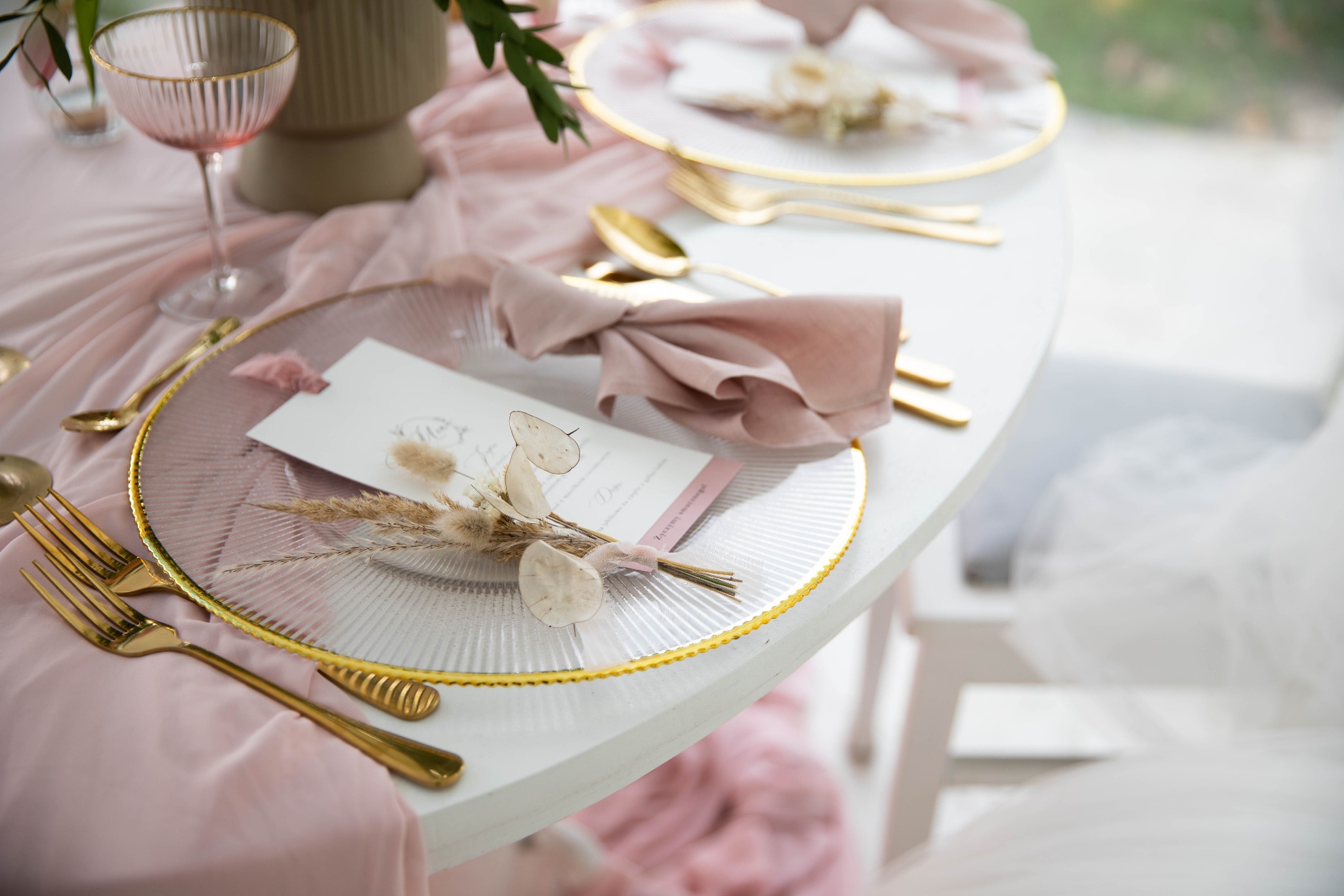 Elegancko nakryty stół w Piasecznie z przezroczystymi talerzami ze złotym obramowaniem, złotymi sztućcami, różową serwetką i kwiatową dekoracją, przygotowany na przyjęcie.