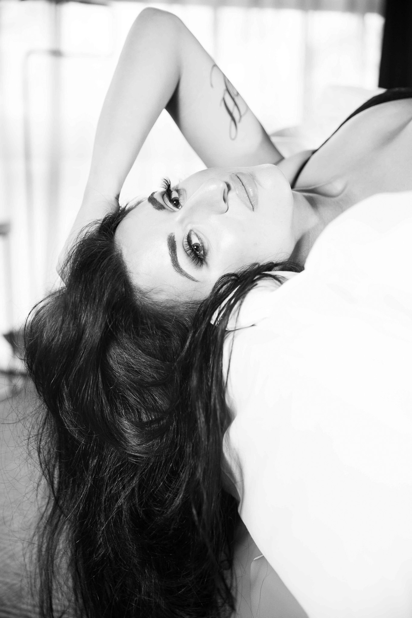 Portret kobiety leżącej na białej pościeli, z tatuażem na ramieniu, w czarno-białej tonacji, z wyraźnym makijażem oczu.