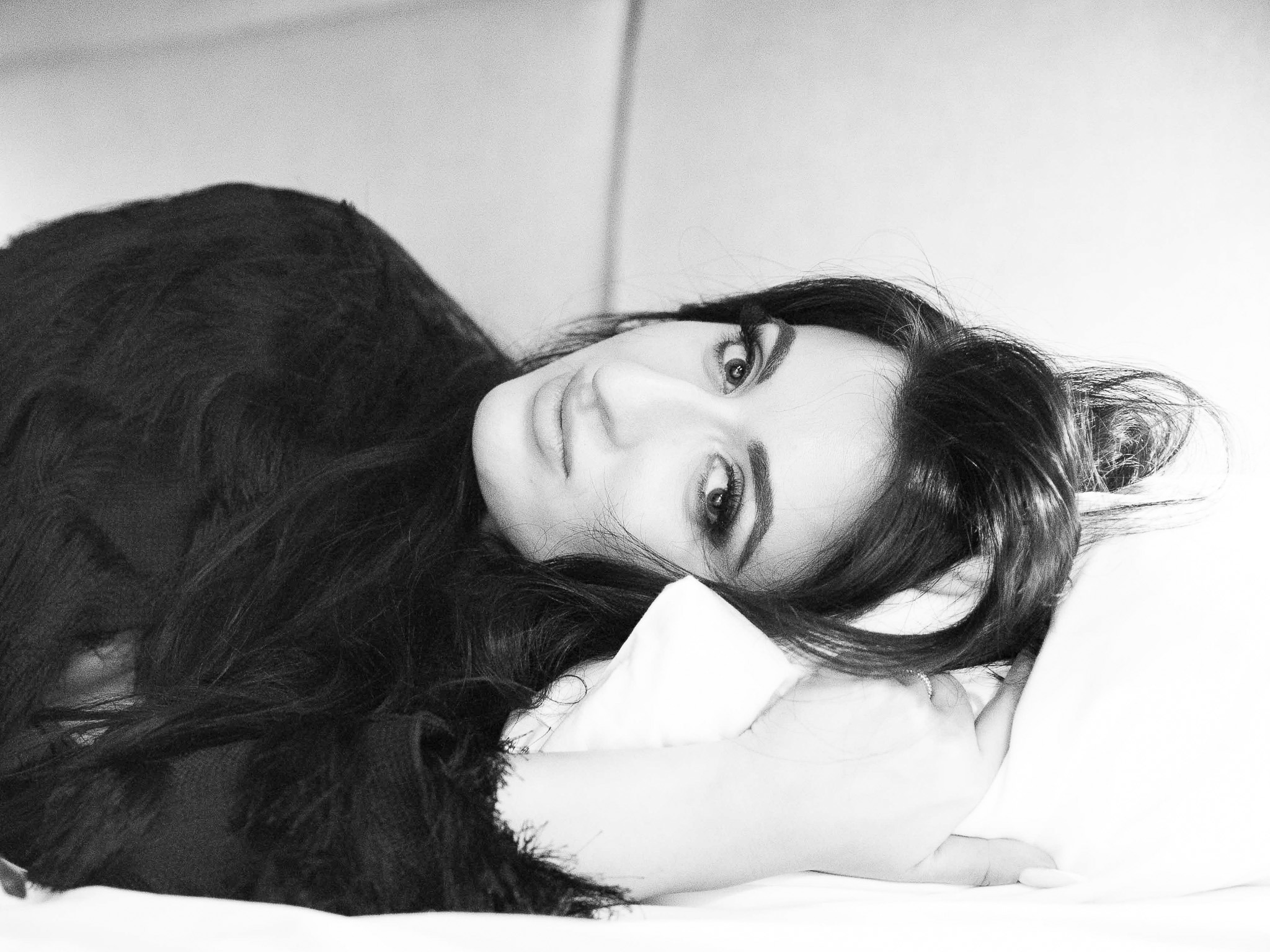 Portret kobiety z długimi, ciemnymi włosami, leżącej na białej poduszce, w czarno-białej tonacji, z intensywnym spojrzeniem w obiektyw.