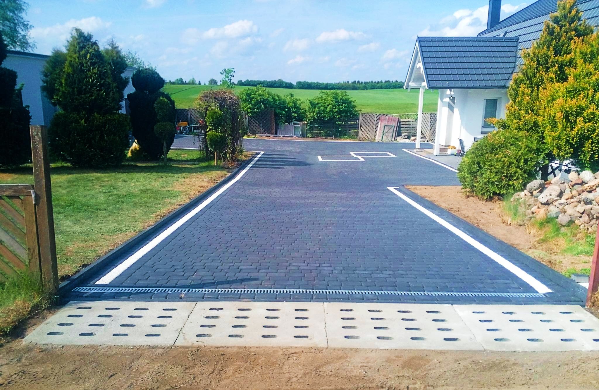 Ułożona, ciemnoszara kostka brukowa z białymi liniami wyznaczającymi miejsca parkingowe na podjeździe w Czersku, zakończona betonowym odpływem liniowym.