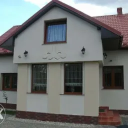 Jasny dom jednorodzinny z czerwonym dachem i oknami w drewnianych ramach, otynkowany na biało, z dekoracyjnym elementem nad wejściem i zewnętrznymi lampami.