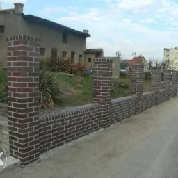 Nowo wybudowany, niski murek z cegły klinkierowej z filarami, oddzielający posesję od drogi. Widoczne budynki w tle.