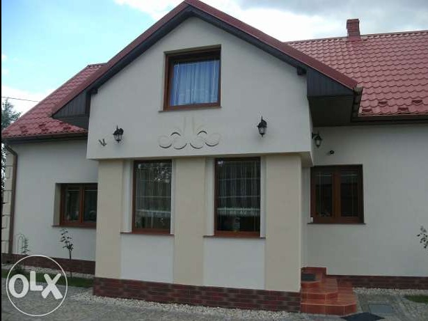 Jasny dom jednorodzinny z czerwonym dachem i oknami w drewnianych ramach, otynkowany na biało, z dekoracyjnym elementem nad wejściem i zewnętrznymi lampami.