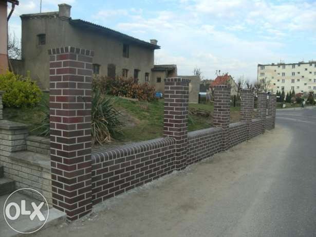 Nowo wybudowany, niski murek z cegły klinkierowej z filarami, oddzielający posesję od drogi. Widoczne budynki w tle.