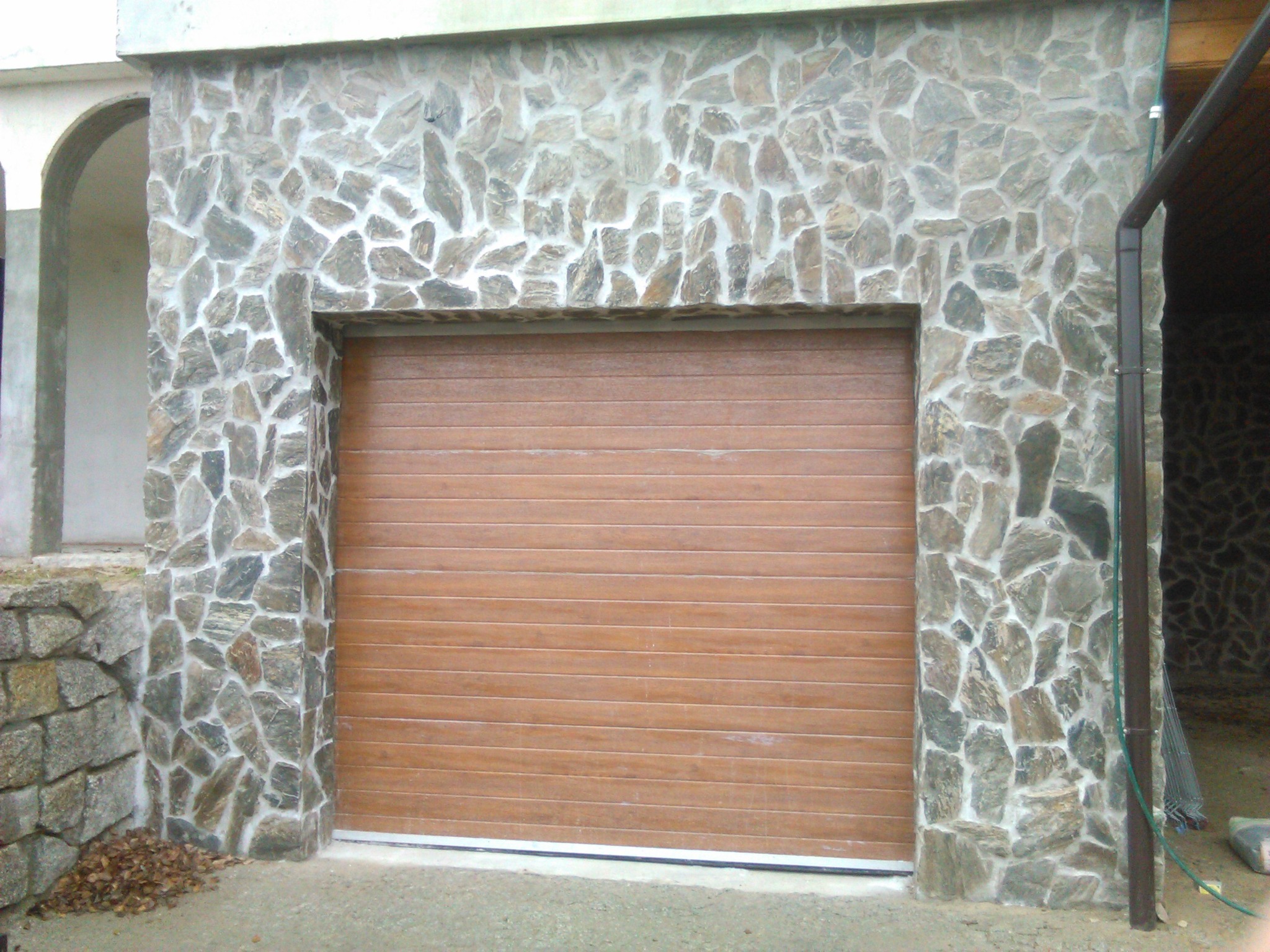 Brama garażowa segmentowa w kolorze drewna, obudowana ścianą z nieregularnego kamienia w odcieniach szarości i beżu, z widocznymi fugami, na zewnątrz budynku.