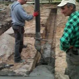 Wylewka fundamentów domu: pracownik w szarej bluzie steruje pompą do betonu, drugi obserwuje postęp prac w wykopie zbrojonym stalą. Budowa w toku, widok z góry.