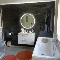 Nowoczesna łazienka z czarnymi marmurowymi płytkami, okrągłym lustrem z podświetleniem LED, białą wiszącą szafką z umywalką i wanną z szarą poduszką. Na podłodze dywaniki w odcieniach pomarańczy.