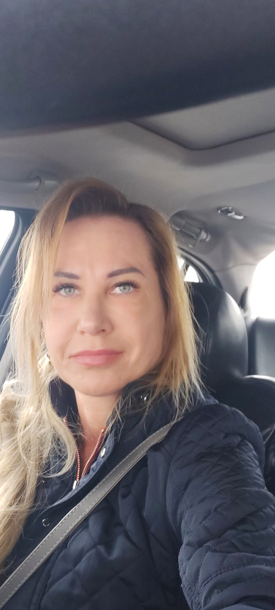 Portret kobiety z blond włosami i niebieskimi oczami, siedzącej w samochodzie, ubrana w pikowaną kurtkę.