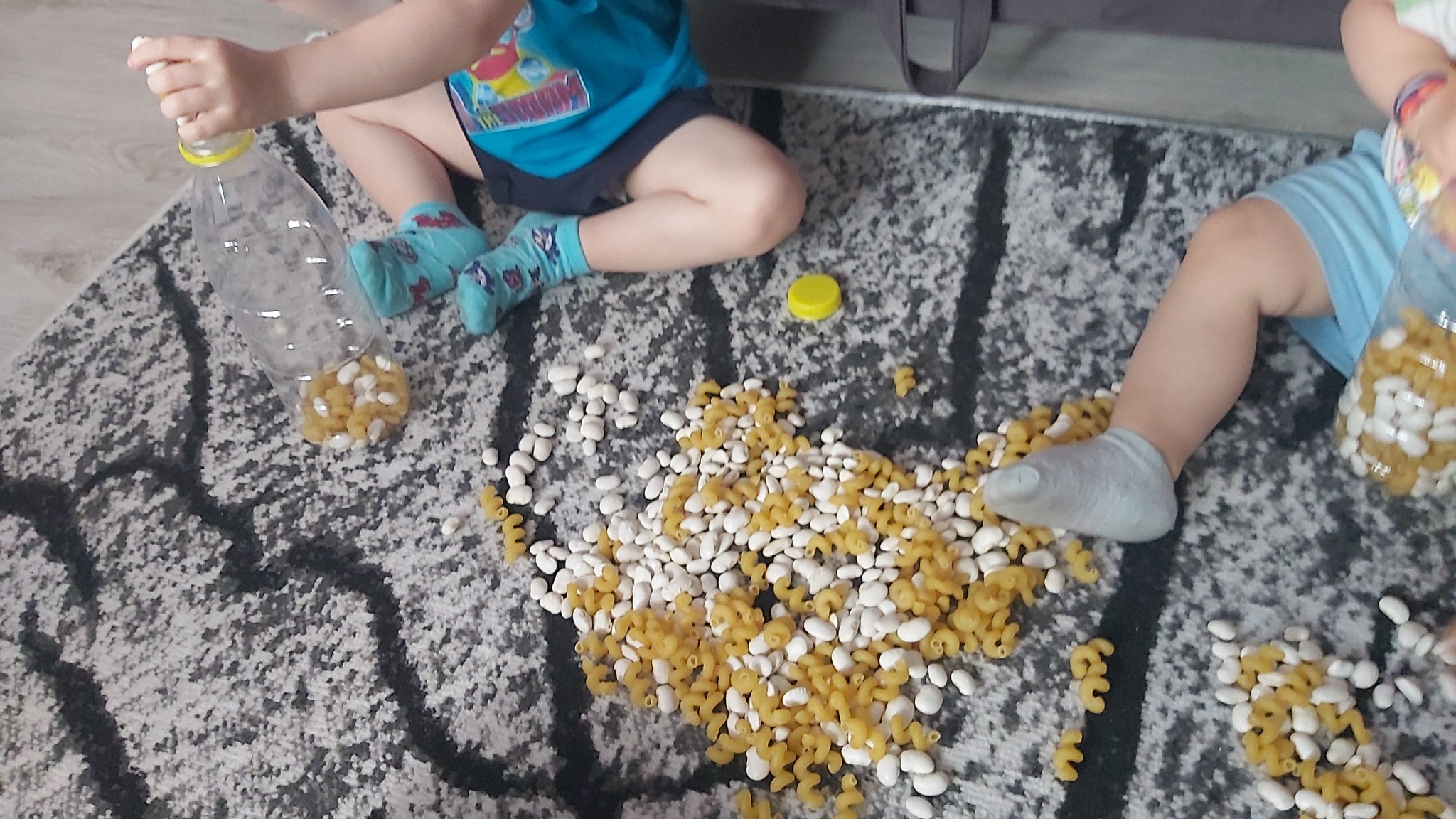 Dzieci bawią się na dywanie, przesypując białą fasolę i makaron świderki do plastikowych butelek. Jedno dziecko ma na sobie niebieskie skarpetki w dinozaury.