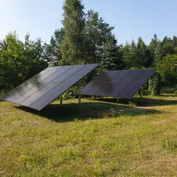 Dwa panele słoneczne na metalowych stelażach ustawione na trawniku przed domem, w tle wysokie drzewa i błękitne niebo, w trawie spaceruje mały pies.