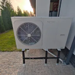 Zewnętrzna jednostka klimatyzacji z logo Netsu, zamontowana na czarnym metalowym stelażu z regulowanymi nóżkami, ustawiona na kostce brukowej przed budynkiem z widocznym systemem odprowadzania...