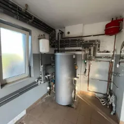 Wnętrze kotłowni z szarym zasobnikiem ciepłej wody, liczne rury i zawory, oraz czerwony zbiornik wyrównawczy na ścianie.