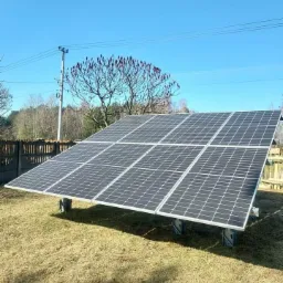 Instalacja paneli słonecznych w Trojanowie na trawiastym terenie, widoczne mocowania, w tle drzewo z czerwonymi kwiatostanami i drewniany płot.