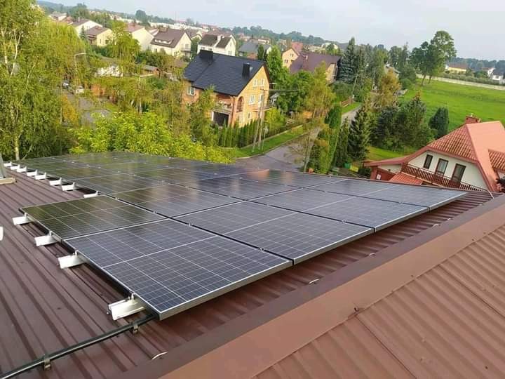 Panele słoneczne zamontowane na brązowym dachu blaszanym, widok na zabudowę jednorodzinną w oddali.