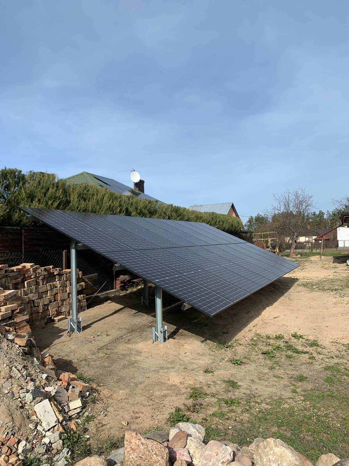 Panele fotowoltaiczne ustawione na metalowych wspornikach na ziemi, z stertą cegieł w tle i domami w oddali.