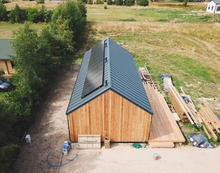 Widok z góry na dom z drewnianą elewacją i panelami słonecznymi na grafitowym dachu. Część tarasu w budowie, widoczne deski i materiały budowlane.