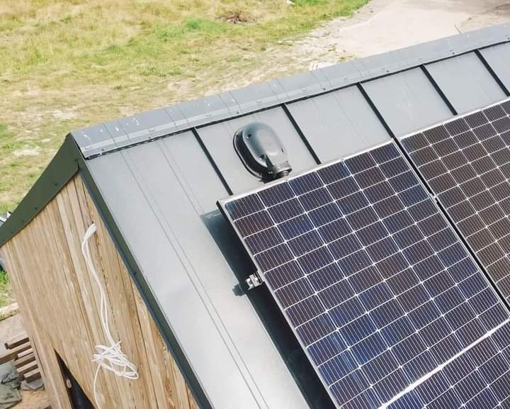 Panele słoneczne na dachu budynku z drewnianą elewacją, widok z góry pod kątem, widoczne kable zwisające ze ściany.