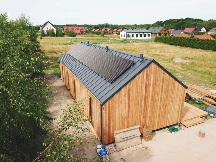 Nowoczesny dom z drewnianą elewacją i panelami słonecznymi na grafitowym dachu, widok z lotu ptaka na tle wiejskiej zabudowy.