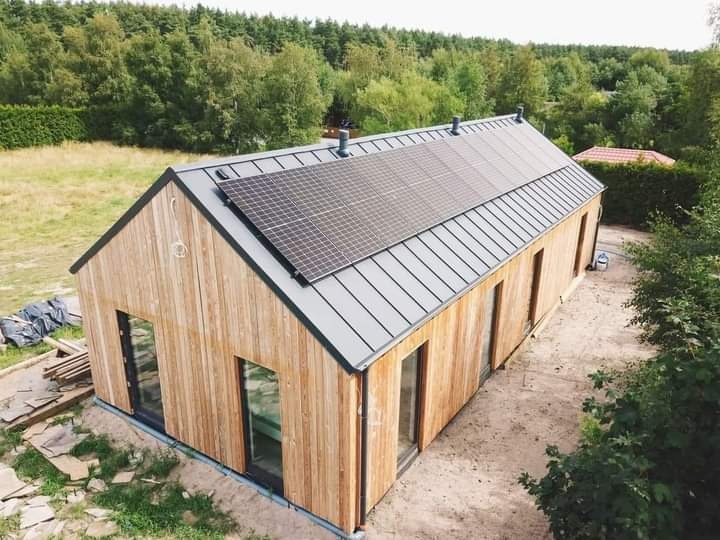 Nowoczesny dom z jasnego drewna z szarym dachem i panelami słonecznymi, widok z góry, teren wokół domu częściowo zagospodarowany.