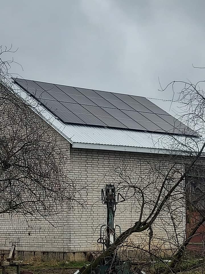 Czarny panel fotowoltaiczny na białym blaszanym dachu ceglanego budynku gospodarczego, widok przez gałęzie drzewa.
