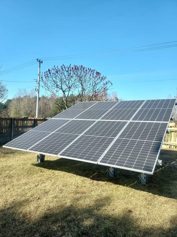 Instalacja paneli słonecznych w Trojanowie na trawiastym terenie, widoczne mocowania, w tle drzewo z czerwonymi kwiatostanami i drewniany płot.