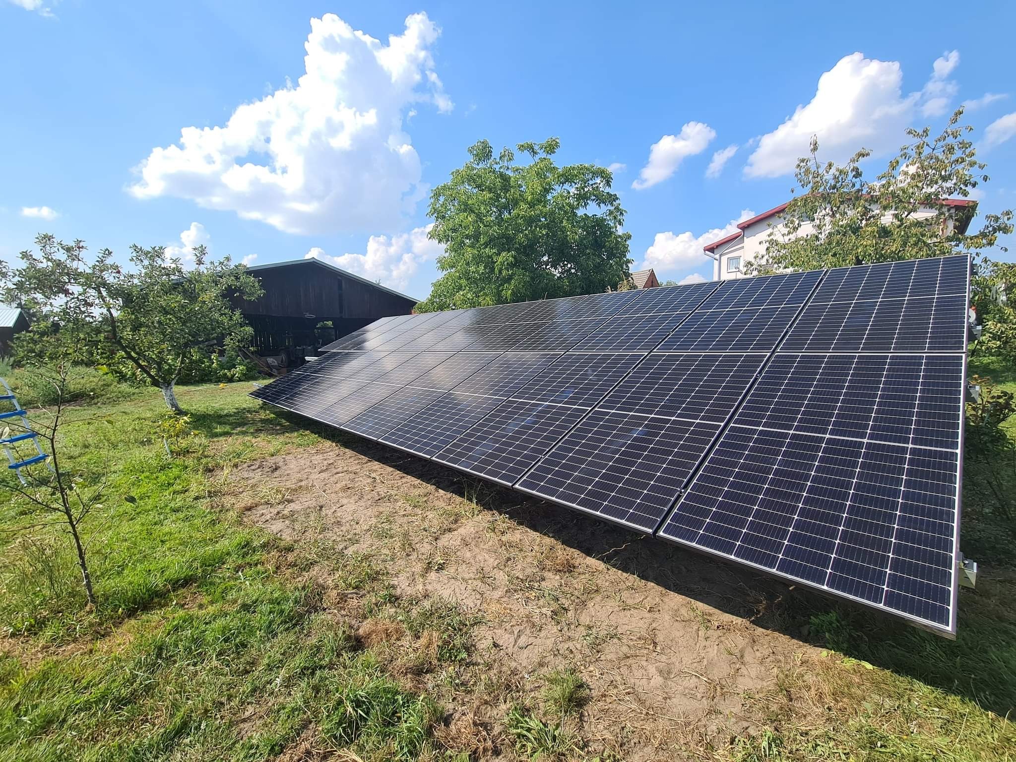 Instalacja paneli słonecznych na gruncie z widocznym budynkiem gospodarczym i domem jednorodzinnym w tle, błękitne niebo z chmurami.