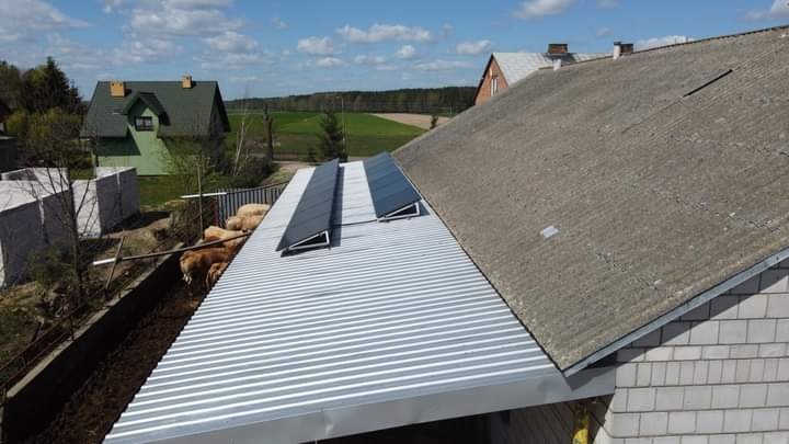 Panele słoneczne zamontowane na blaszanym dachu budynku gospodarczego, obok dachu krytego eternitem, w tle zabudowania wiejskie i pole.