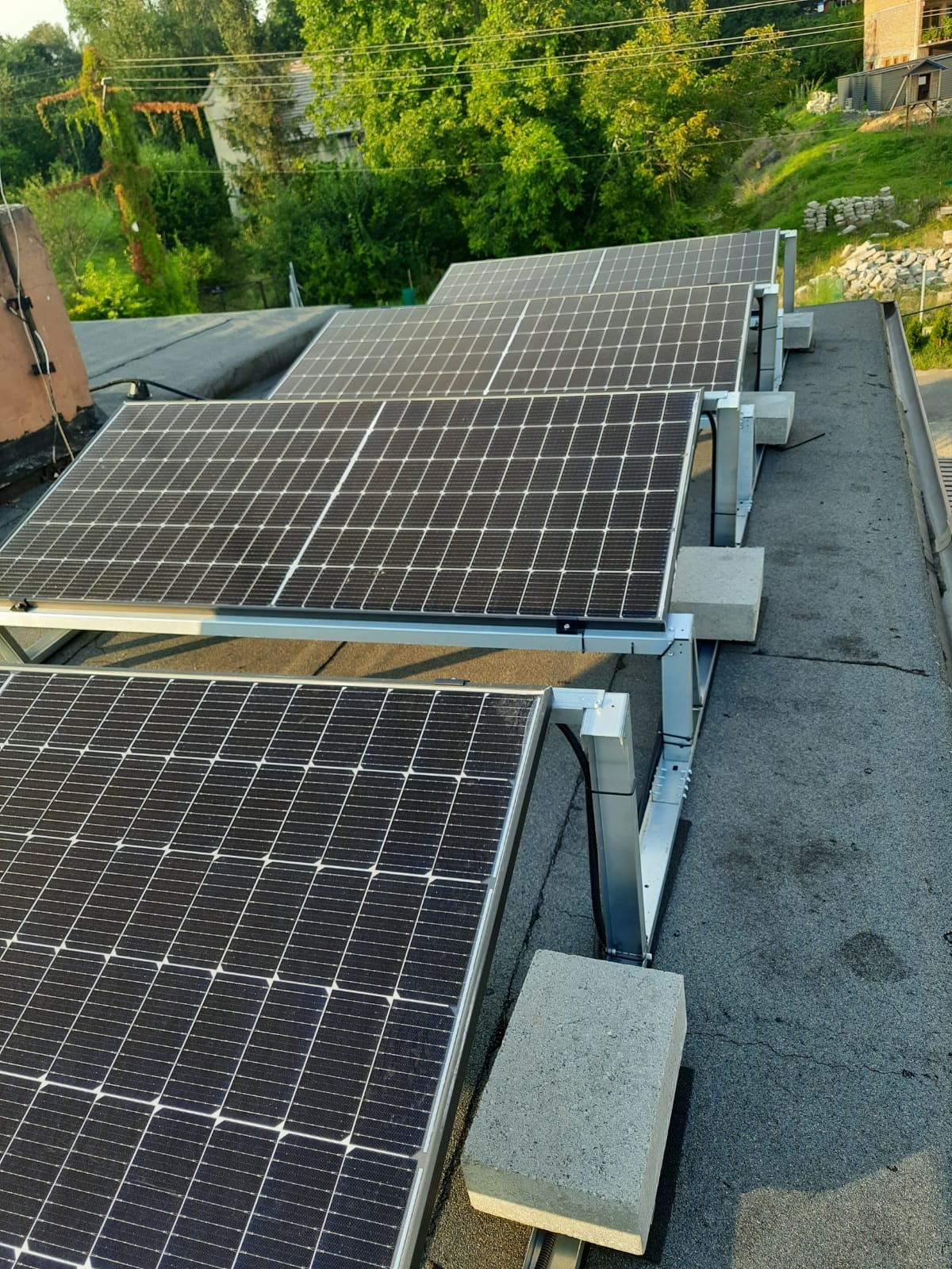 Rząd paneli słonecznych na płaskim dachu, wsparty na metalowych konstrukcjach obciążonych betonowymi blokami, z zielenią w tle.