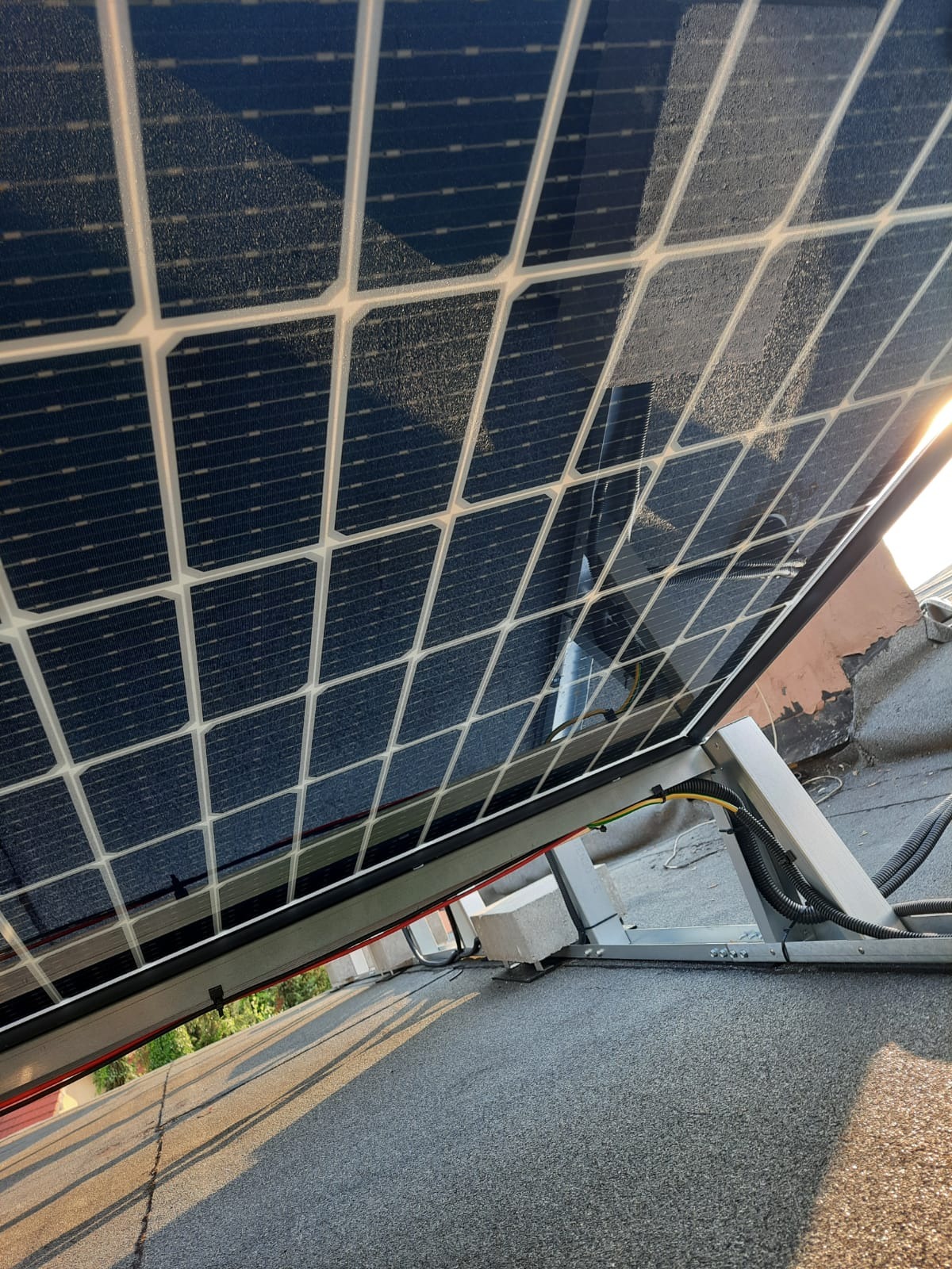 Nisko kątowe ujęcie panelu słonecznego na dachu pokrytym papą, widoczne okablowanie i fragmenty konstrukcji montażowej.