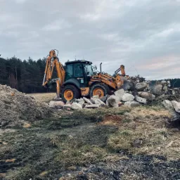 Żółto-czarna koparko-ładowarka Case 695 Super R podczas rozbiórki gruzowiska na zewnątrz, w tle las iglasty pod pochmurnym niebem.