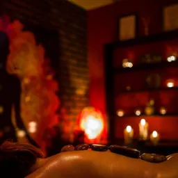 Osoba leżąca na plecach z kamieniami na ciele, w tle dekoracja ściany z motywem sylwetki, półka z dekoracjami i świecami w przyciemnionym, ciepłym świetle.