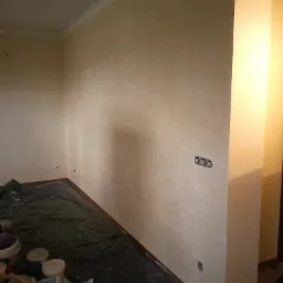 Pokój w trakcie malowania ścian na beżowo, widoczne zabezpieczone folią podłoga i puszki z farbą.