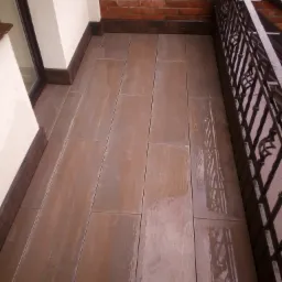 Wykończony balkon z brązowymi płytkami imitującymi drewno, z metalową balustradą i ceglaną ścianą w tle.