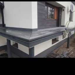 Wykończony taras przydomowy z szarą, kompozytową deską tarasową, widoczna antena satelitarna na ścianie budynku.