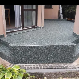 Wykończony taras z żywicy epoksydowej w odcieniach szarości, z widocznymi schodami wejściowymi, kolumną i fragmentem ogrodu z roślinnością.