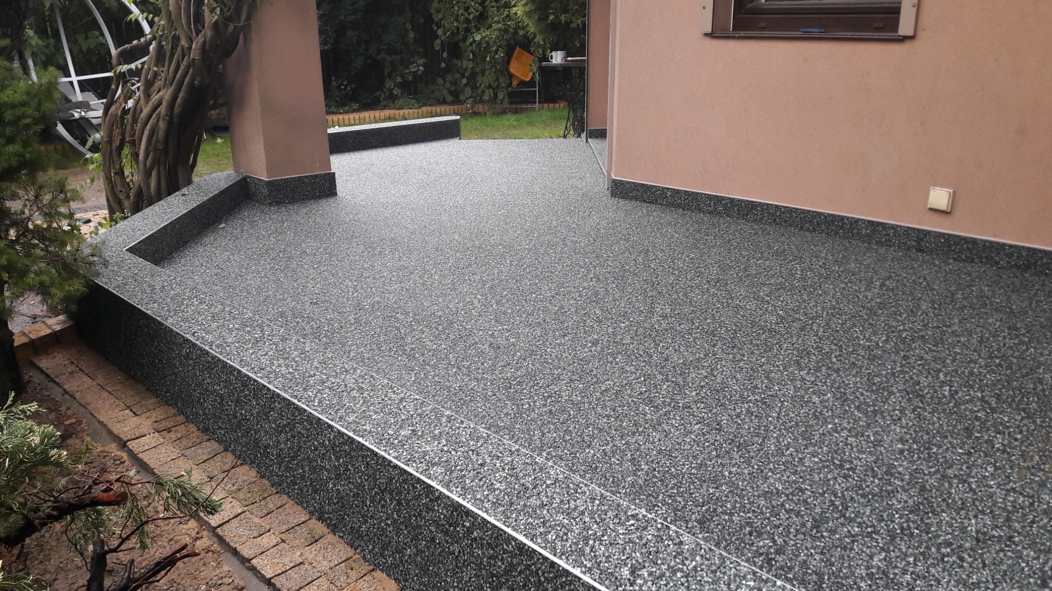 Wykończony taras z szarą, żywiczną posadzką z drobnymi kamyczkami, obrzeża wykończone listwą, widoczne schodki i fragment elewacji budynku w tle zieleń ogrodu.