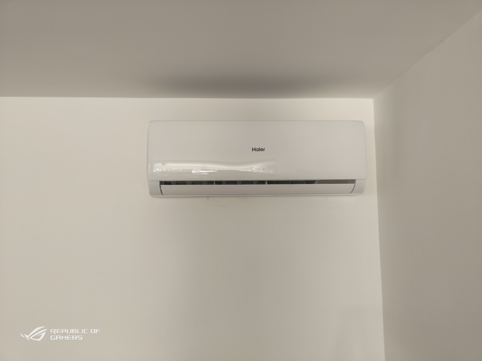 Biała jednostka klimatyzacyjna Haier zamontowana na białej ścianie w rogu pokoju, widoczny fragment logo Republic of Gamers w lewym dolnym rogu.