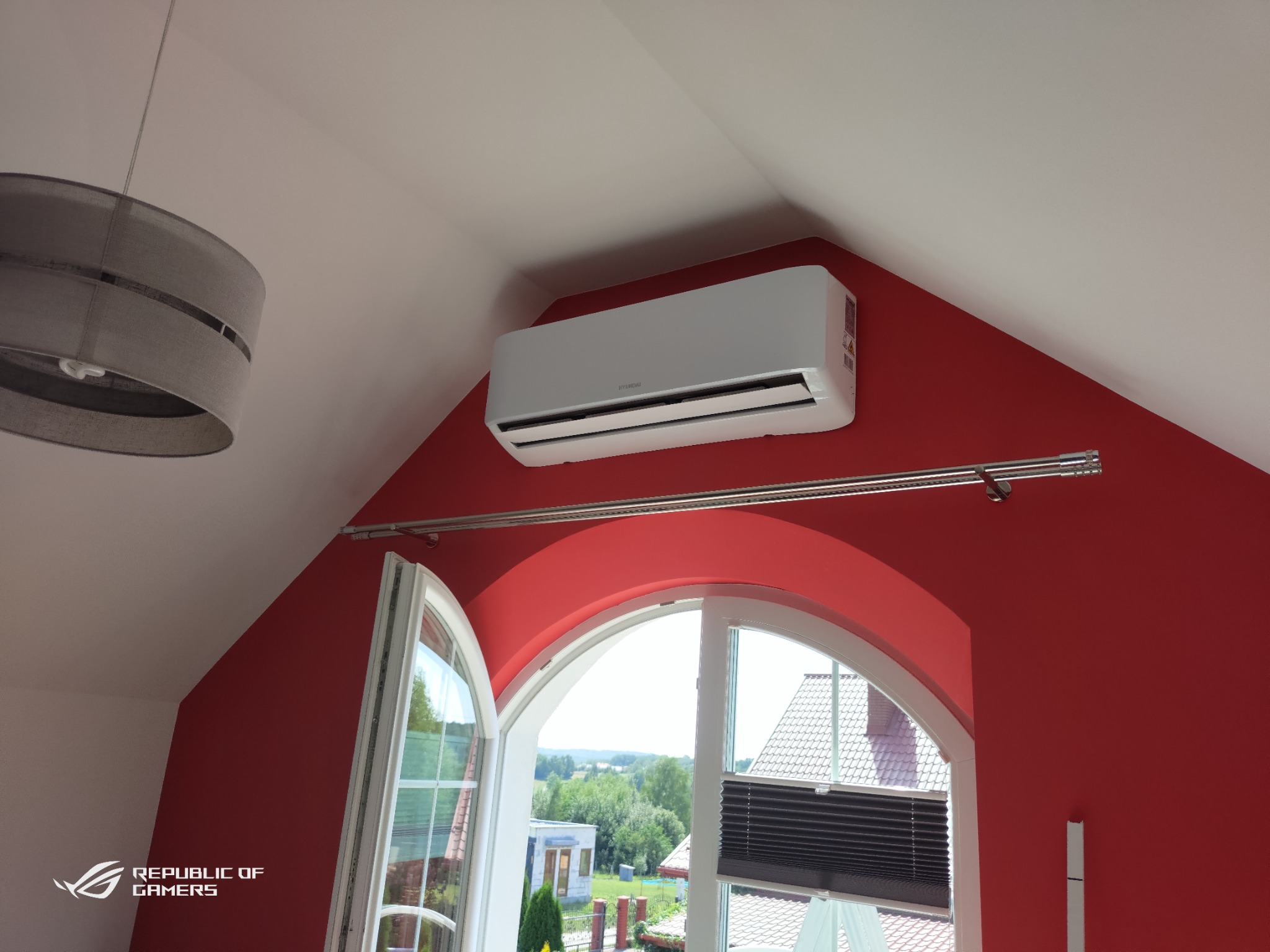 Biały klimatyzator Hyundai zamontowany na czerwonej ścianie pod skosem, widok z dołu, okno łukowe z widokiem na zieleń w tle.
