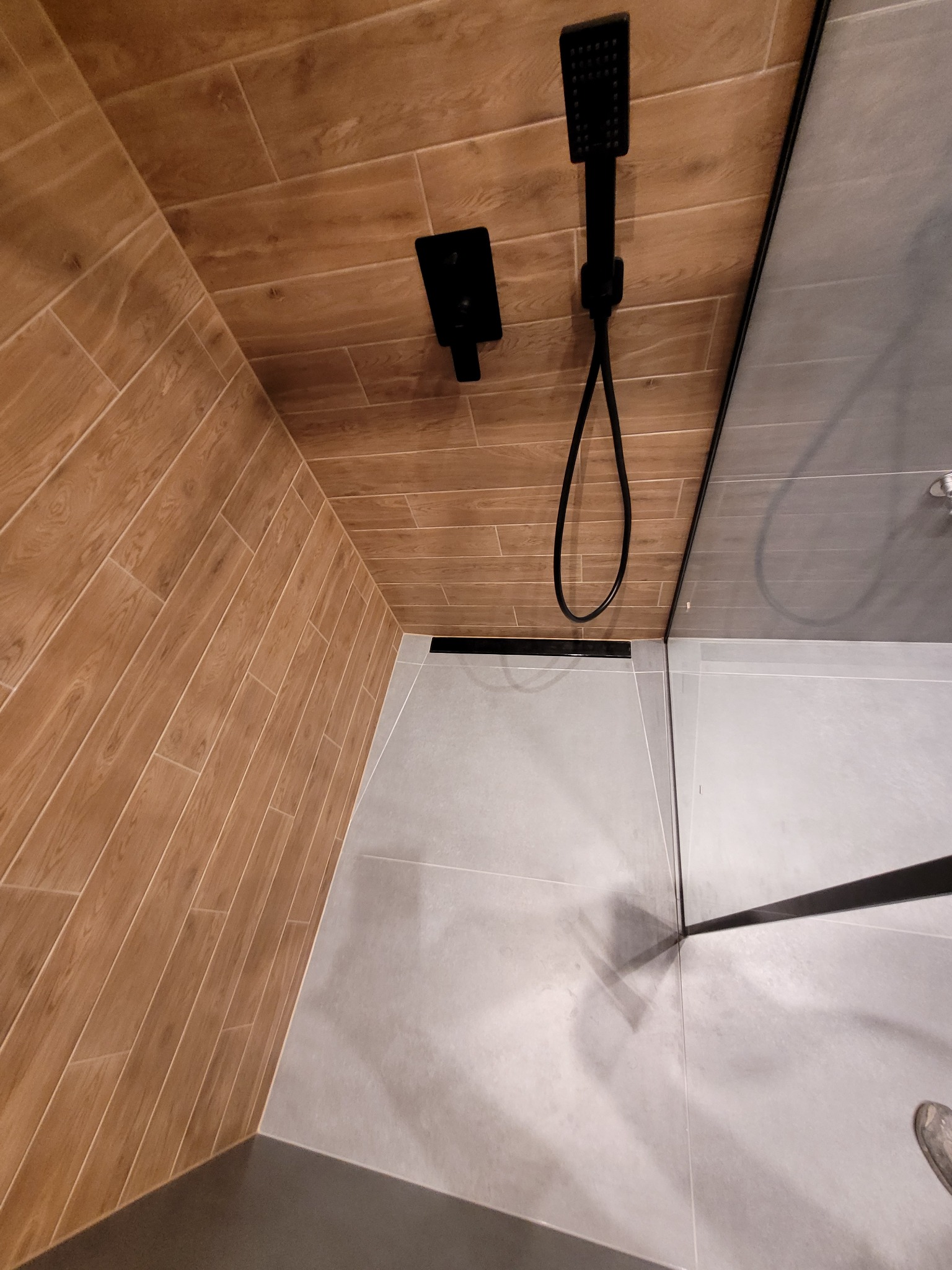 Nowoczesna kabina prysznicowa z czarną armaturą, wykończona płytkami imitującymi drewno na ścianie i szarymi płytkami na podłodze, z odpływem liniowym.