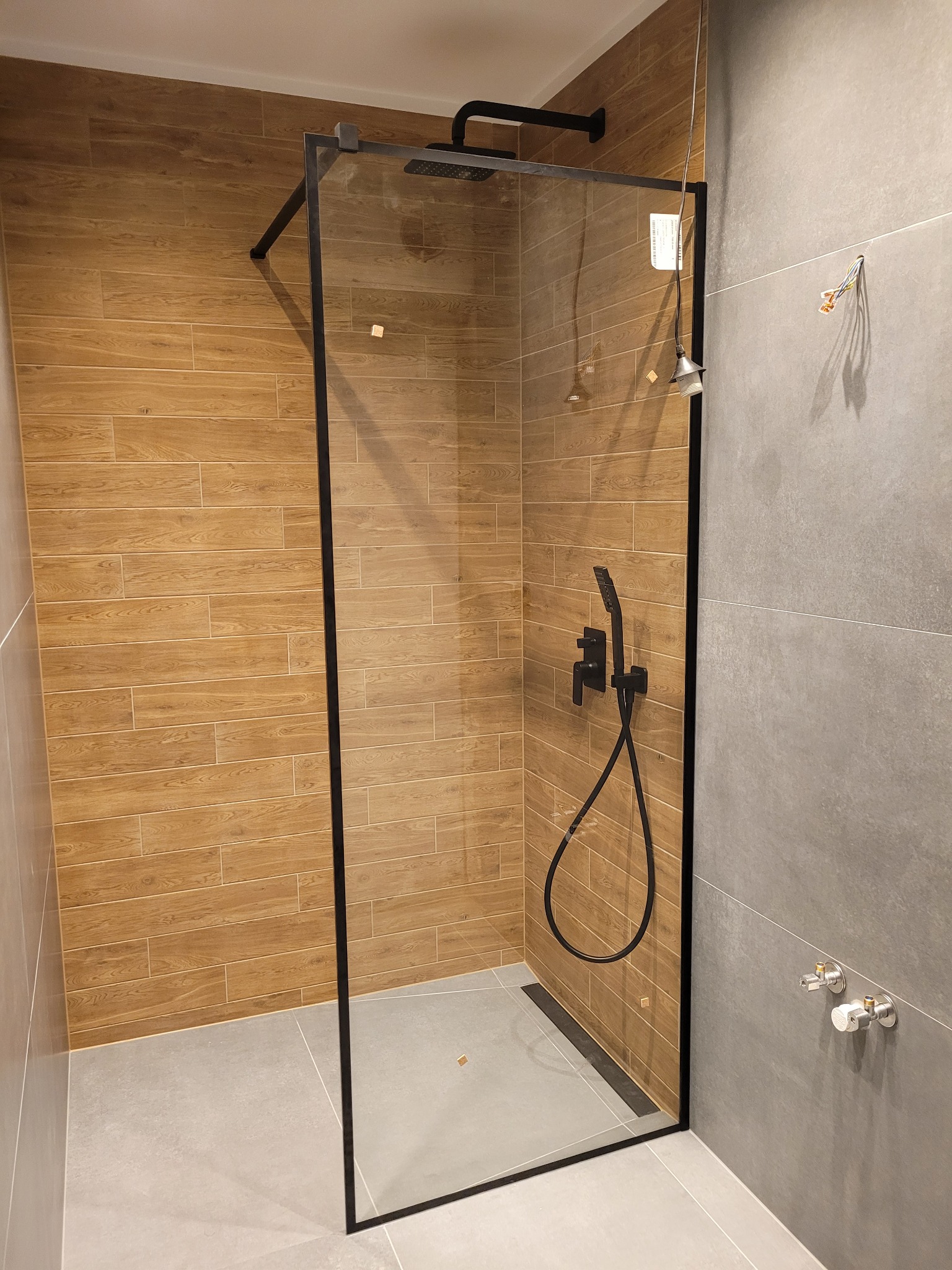 Nowoczesna kabina prysznicowa z czarną ramą i armaturą, wykończona płytkami imitującymi drewno i szarymi płytkami wielkoformatowymi, z widocznymi przyłączami hydraulicznymi.