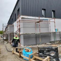 Budynek w trakcie wykańczania elewacji, widoczne rusztowania, pracownik w ubraniu roboczym, na ziemi materiały budowlane i śmieci.