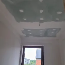 Niedokończony sufit z płyt kartonowo-gipsowych, widoczne ślady szpachlowania i otwory na wkręty, w tle okno z widokiem na budynek w trakcie budowy.