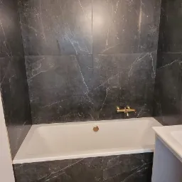 Wykończona łazienka z wanną obudowaną czarnymi płytkami imitującymi marmur, złotą armaturą i drewnopodobną podłogą. Widoczne wystające przewody elektryczne.